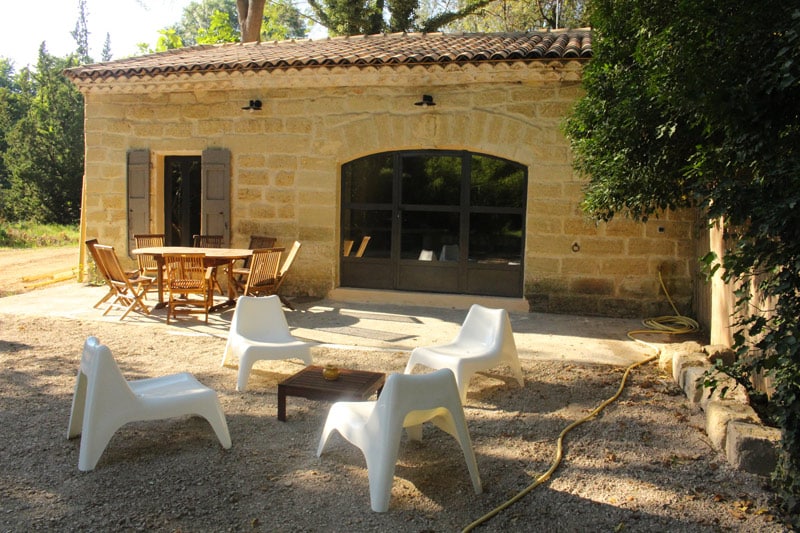 Maison en pierre, terrasse avec salon de jardin en bois et fauteuils blancs design au soleil.